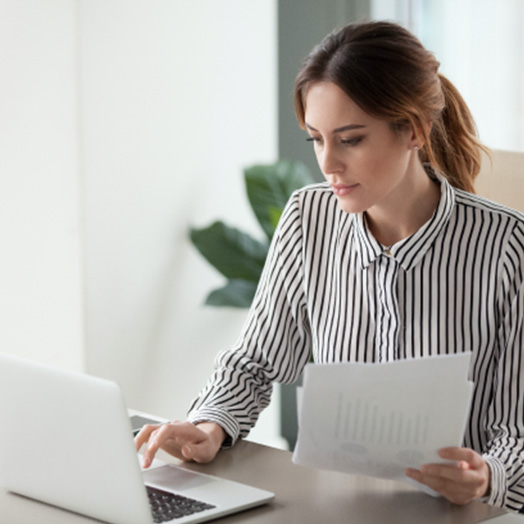 A young professional women typing on a laptop and holding financial documents.