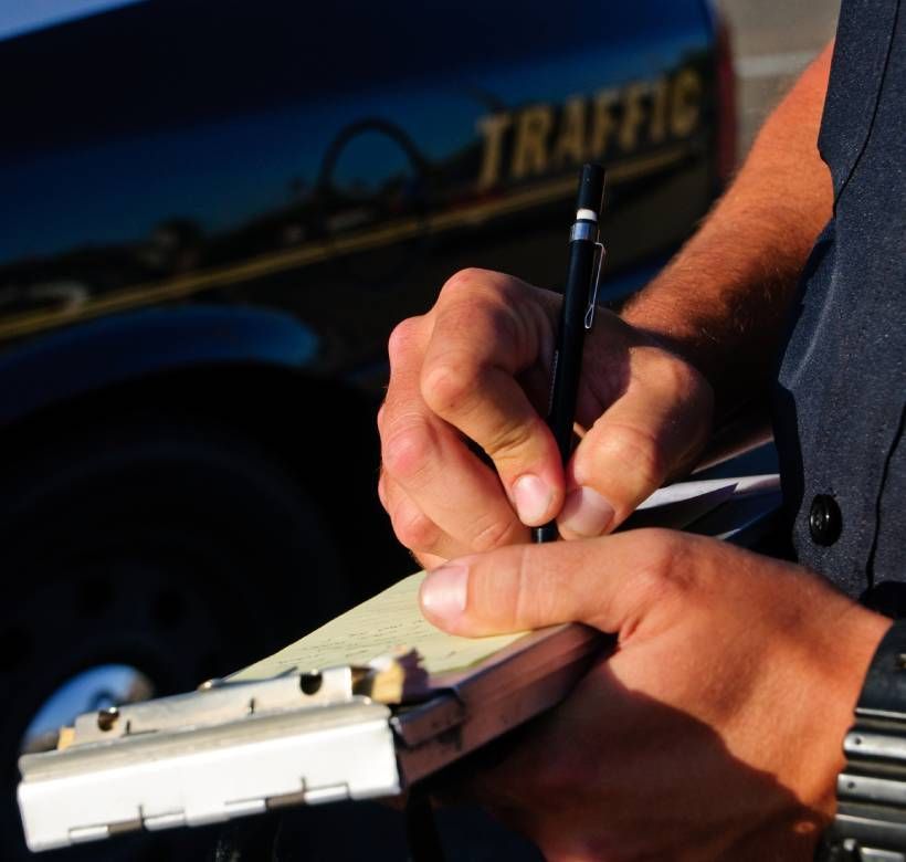 Policía de tráfico escribiendo una multa por infracción de tráfico en movimiento mientras permanece de pie junto a su vehículo patrulla.