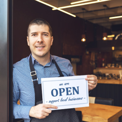 A business owner standing in the doorway of his restaurant with a sign that reads, we are OPEN. support local businesses.